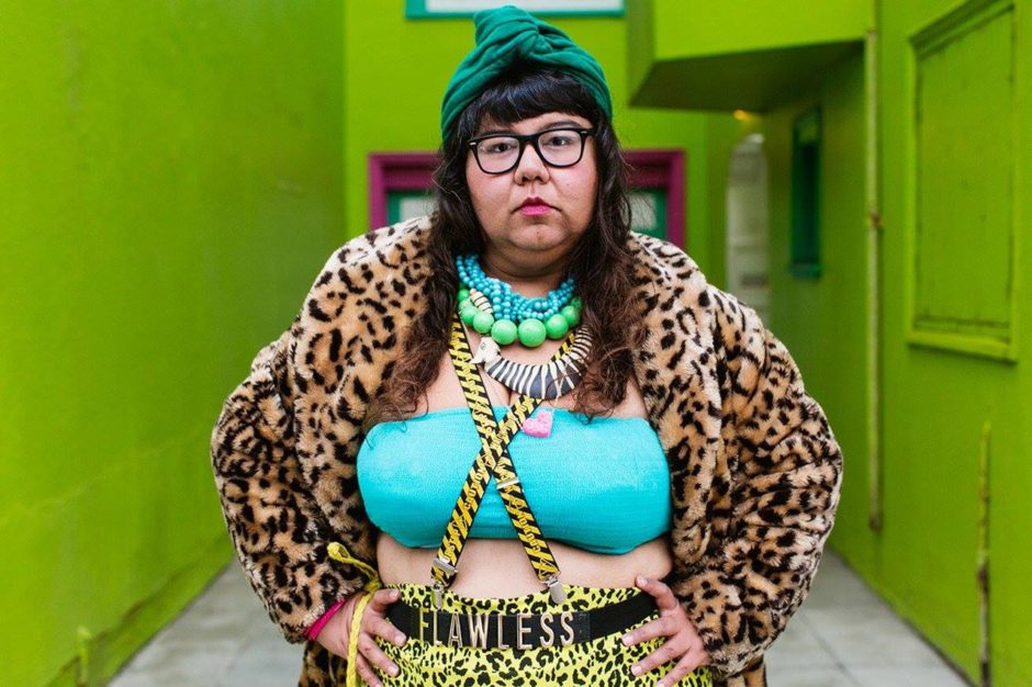 Virgie stares at the viewer with a green background, her bra. hat and jewelry are teal. Her coat and pants are animal print. In the middle of this dream is a belt buckle reading "FLAWLESS"
